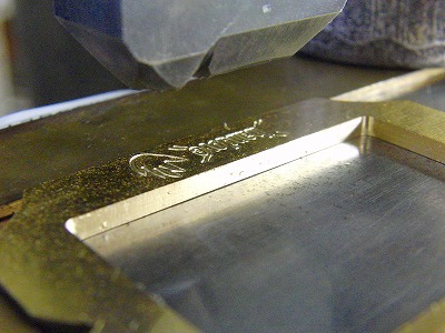 真ちゅうを彫刻機にて削り出し、成形した後に、お客様ご指定のロゴとモチーフを手作業で刻印していきます。  上部に少し見えるのが鋼製（日本刀と同じ材料）の刻印になります。